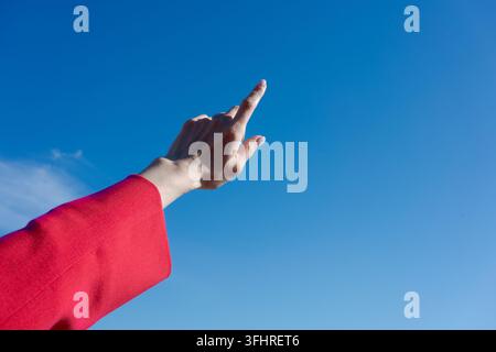 Hand pointing up towards the sky. The sky is blue and clear. The hand is wearing a red jacket Stock Photo