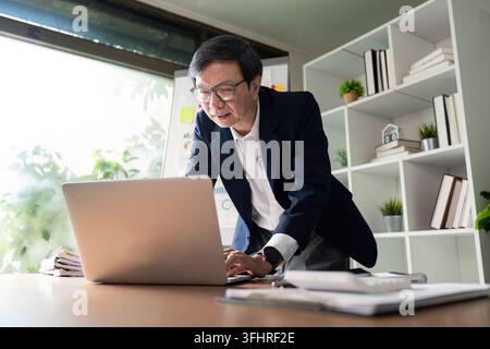 An elderly businessman focused on his laptop, analyzing data and preparing for financial management, surrounded by documents and a modern office envir Stock Photo