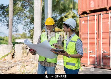 Two engineers in safety helmets and vests are smiling while reviewing logistics plans and collaborating at a shipping container site, showcasing teamw Stock Photo