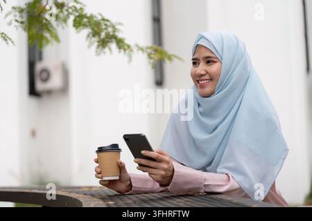 Muslim business woman in hijab smiling while holding coffee and smartphone. Stock Photo