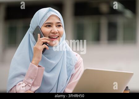 Smiling Muslim businesswoman in hijab talking on the phone while working on a laptop outdoors. Stock Photo