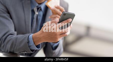Close up of a young Asian businessman holding a smartphone and a snack in a modern office environment. Stock Photo