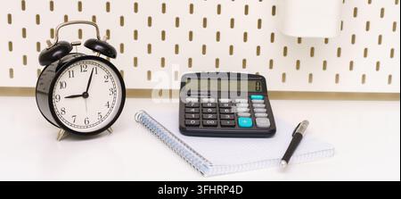 The calculator lies on notebook against  background of an alarm clock on table. Stock Photo