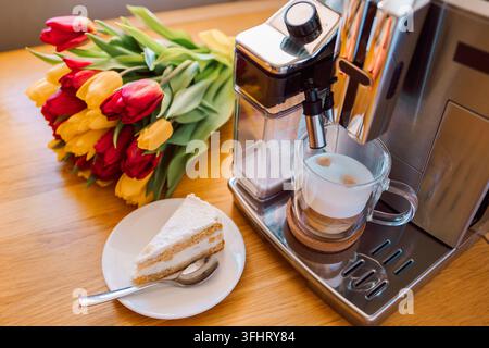 Cup of coffee with milk, piece of cake and tulips flowers on wooden kitchen table. Freshly brewed cappuccino espresso in coffee Stock Photo