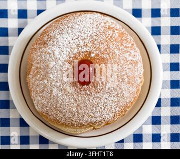Top down view of a traditional Hanukkah doughnut with red jam and powdered sugar on a white plate over a blue and white checkered tablecloth. Stock Photo