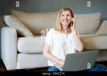 Beautiful adult woman is sitting on floor by sofa and talking and laughing Stock Photo