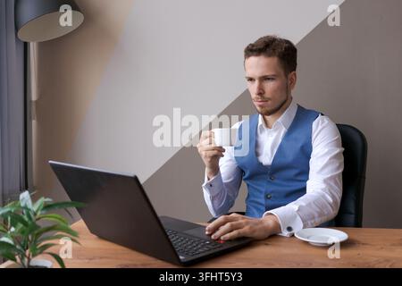 Businessman is working on laptop for new architectural project. Caucasian young man in a shirt and waistcoat sits at a table in the office and works. Stock Photo