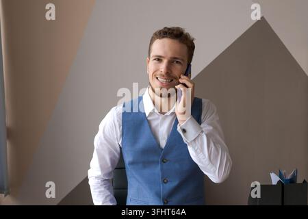 Pleasant handsome bearded man in shirt and waistcoat talking on phone, ordering taxi or food delivery to office at work. Smiling young caucasian busin Stock Photo