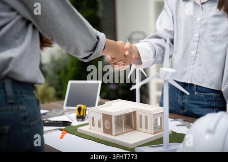 Two sustainability engineers shaking hands as a sign of agreement on their project, with architectural models and blueprints on the table outdoors. Stock Photo