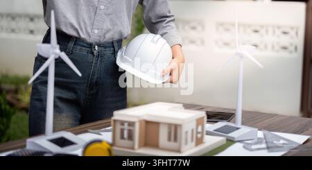 An engineer holding a helmet examines a sustainable architectural model featuring wind turbines, showcasing innovation in eco-friendly building design Stock Photo
