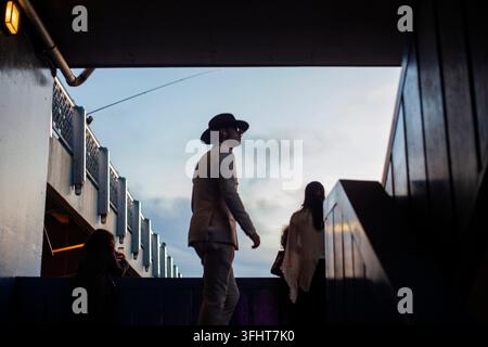 A young man wearing vintage-inspired clothing and a wide-brimmed hat walks across Galata Bridge in Istanbul, Stock Photo