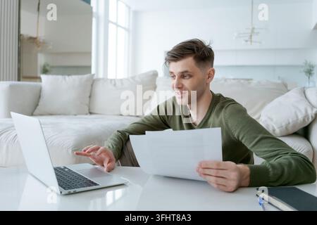 Man is sitting on the floor in front of a laptop, looking at a piece of paper. He is holding the paper in his hand and he is focused on it Stock Photo