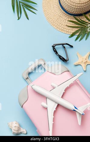 Pink suitcase with toy airplane, sunglasses, straw hat, seashells and palm leaves on blue background Stock Photo