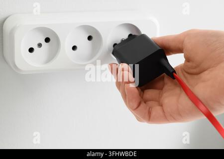 Energy consumption and power supply, hand plugging a black charger into a white wall socket for electricity and device charging in a home office Stock Photo