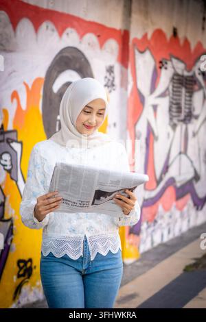 A young Asian Muslim woman is reading a newspaper by the roadside. Stock Photo