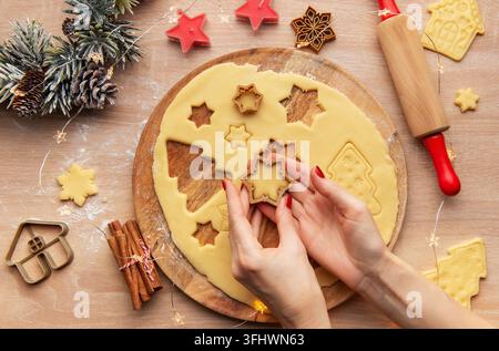 Woman preparing christmas cookies using different shaped cookie cutters on a wooden board, surrounded by festive decorations Stock Photo