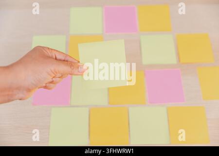 Hand holding a blank green sticky note above organized colorful notes on a wooden desk, representing selection, priority, planning, task management, d Stock Photo
