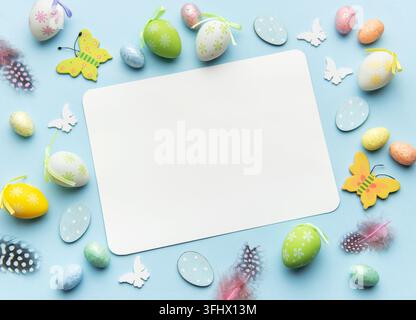 Easter eggs, feathers and butterflies forming frame around blank card on blue background Stock Photo