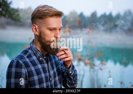 The bearded man smokes. Handsome stylish man in plaid shirt with cigarette. Stock Photo