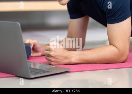 A dedicated young man performs a plank exercise at home, using a laptop for guidance. Focus on fitness and online training. Stock Photo