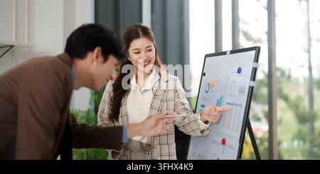 Two colleagues analyze data and trends on a presentation board, collaborating on business strategies in a bright office. Stock Photo
