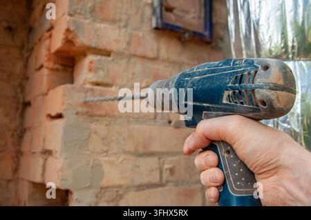 Close-up of a person's hand operating a blue power drill, drilling into a weathered brick wall with a metal bit Stock Photo