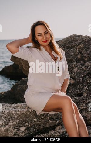 Woman coast sea sitting on rocks by the shore during sunset enjoying a peaceful moment of relaxation Stock Photo