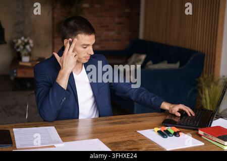 Young man in jacket working from home doing paperwork using laptop and holding a pen in his hand while sitting at his desk. Performs work remotely Stock Photo