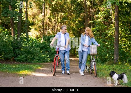 Active old age, people and lifestyle concept happy senior couple fixing bike, talking on summer city park on path in sunny forest, wearing casual clot Stock Photo