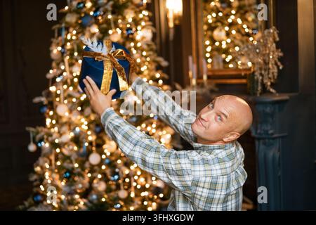 Happy bald man holding a box with a gift on the background of a christmas tree Stock Photo