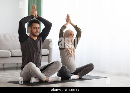 Pregnant Muslim Couple Practicing Yoga At Home, Sitting With Hands Over Head And Closed Eyes On Mat In Living Room, Young Expectant Islamic Spouses Me Stock Photo