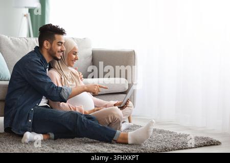Happy islamic family man and pregnant woman using laptop together at home, cheerful middle eastern spouses making online shopping for future baby whil Stock Photo