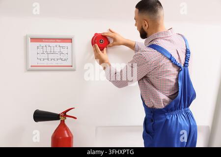 Fire safety inspection and building maintenance, technician installing a fire alarm bell on a wall with an evacuation plan and fire extinguisher nearb Stock Photo