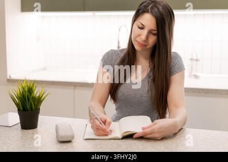 A woman is writing in a notebook on a kitchen counter Stock Photo