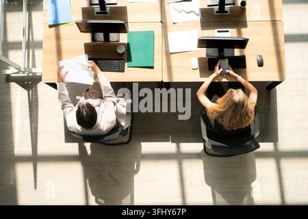 Top view of two professionals working at desks, reviewing documents and using a smartphone in a modern office workspace Stock Photo