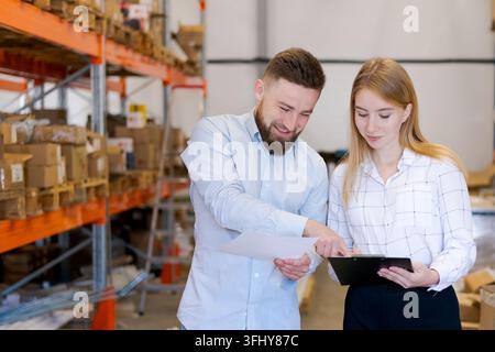 Smiling man and woman logistics supervisors discussing paperwork and pointing at a clipboard in a warehouse. Concept of successful teamwork, distribut Stock Photo