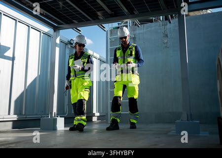 Male and female engineers inspecting photovoltaic systems beneath solar panel structures on a rooftop, representing professional renewable energy mana Stock Photo