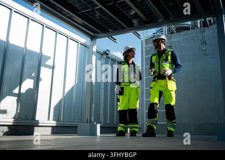 Male and female engineers inspecting photovoltaic systems beneath solar panel structures on a rooftop, representing professional renewable energy mana Stock Photo
