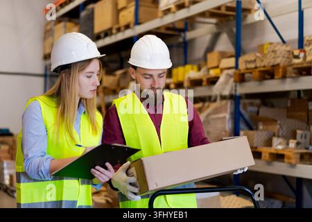 Focused male and female workers examining package details while recording inventory data on clipboard. Meticulous quality control and verification pro Stock Photo