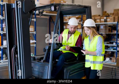 Female logistics manager with clipboard giving instructions to a male forklift operator. Team in safety gear managing stock and distribution in a mode Stock Photo