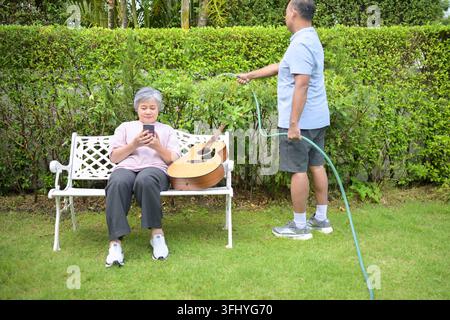 Senior couple spending time in their home garden. Woman using a laptop on a bench while the man sweeps the lawn, representing a balanced and happy ret Stock Photo