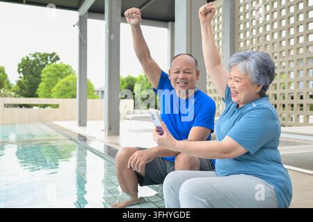 Senior couple sitting by a swimming pool, raising hands in excitement while using a smartphone, representing communication and joyful moments Stock Photo