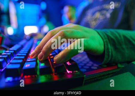 Close-up of a gamer's hands typing on an RGB mechanical keyboard in a dim, colorful gaming setup. Vibrant green and blue lighting highlights keys and Stock Photo