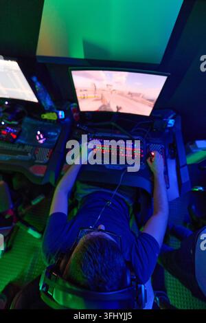 Young man focused on a fast-paced first-person shooter at a neon-lit PC gaming station, using headset, RGB keyboard and mouse in a dark competitive LA Stock Photo