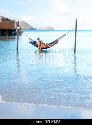A couple unwinds in a hammock gently swaying above crystal-clear water, surrounded by the lush beauty of Saint Lucia. An idyllic setting for a romanti Stock Photo
