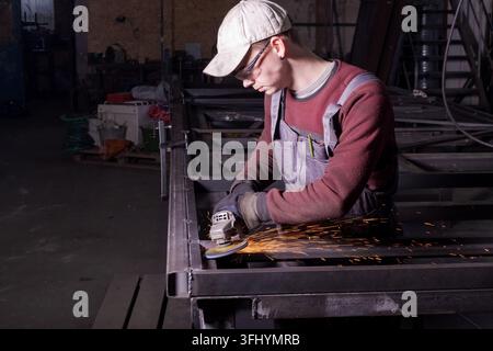 Young man in safety glasses uses angle grinder on steel construction at workshop Stock Photo