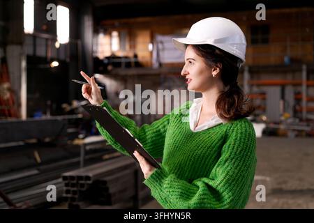 Woman engineer in hard hat points while holding clipboard at metal storage facility Stock Photo