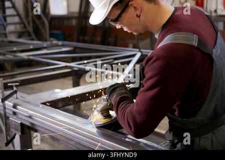 Focused young worker wearing safety helmet using grinder on metal frame with sparks flying in factory Stock Photo