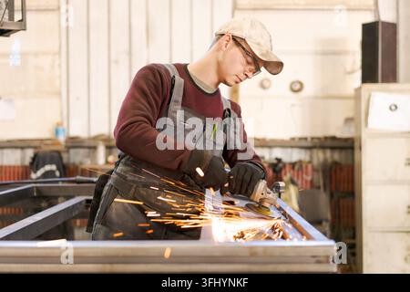 Young man in overalls and cap using power tool on metal construction with flying sparks in manufacturing facility Stock Photo