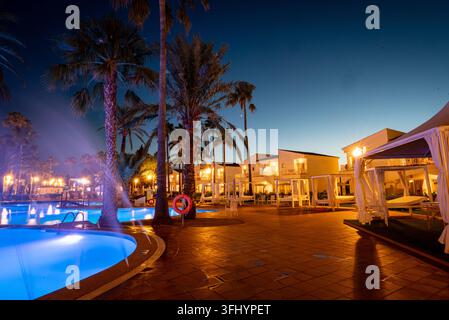 Luxury tropical resort at dusk with glowing swimming pool, palm trees, cabanas and warm lights reflecting on a wet tiled terrace. Stock Photo